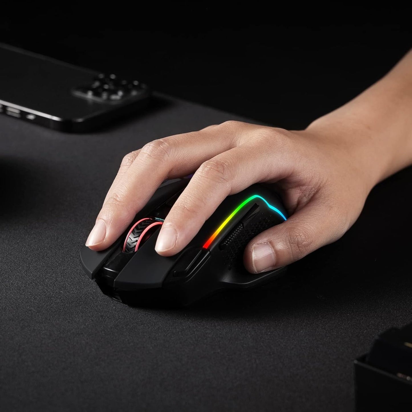 Hand, die eine Gaming-Maus mit RGB-Beleuchtung auf einer dunklen Oberfläche benutzt, mit einem Laptop im Hintergrund.