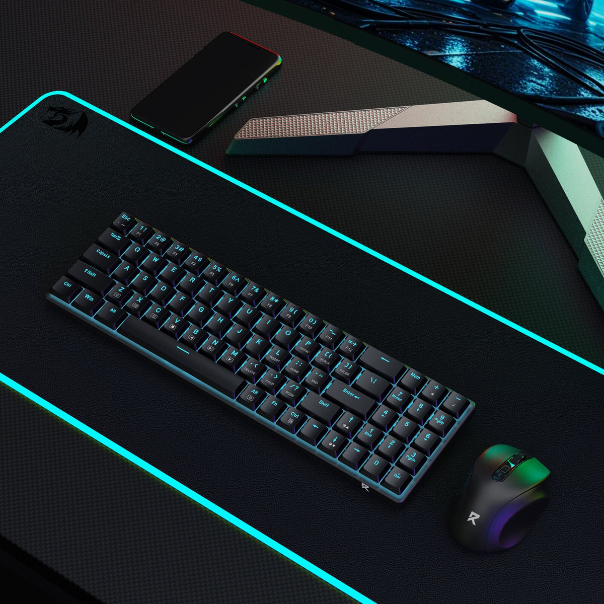 The Redragon keyboard and mouse set is integrated into a gaming setup, placed on an RGB-lit desk mat alongside a monitor and smartphone.
