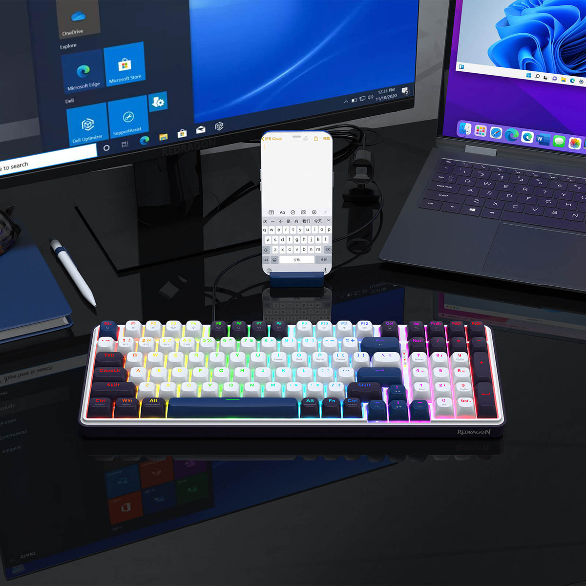 A RGB-lit keyboard (with white and dark keycaps) is placed on a reflective desk, paired with a monitor, laptop, and phone—showcasing its compatibility in a multi-device setup.