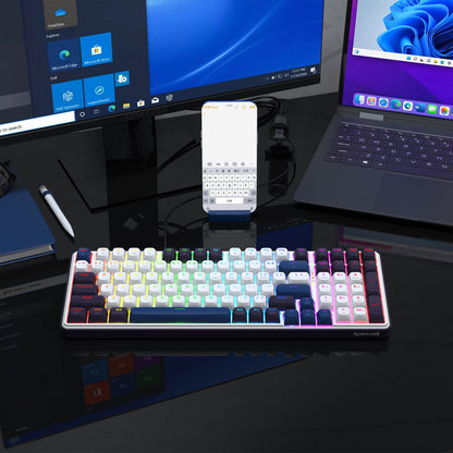 A RGB-lit keyboard (with white and dark keycaps) is placed on a reflective desk, paired with a monitor, laptop, and phone—showcasing its compatibility in a multi-device setup.