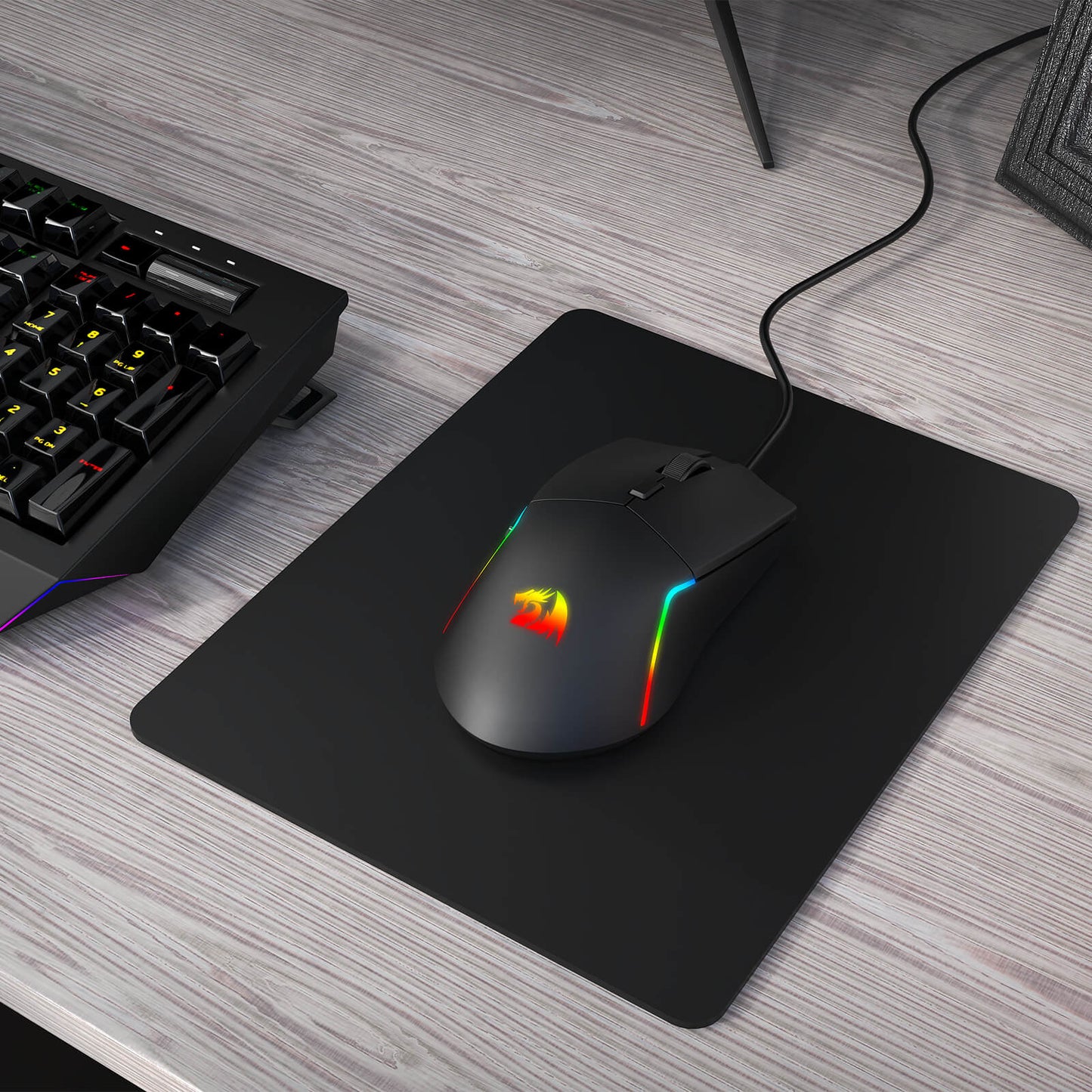 Gaming mouse with RGB lighting on a black mouse pad next to a keyboard on a wooden desk.