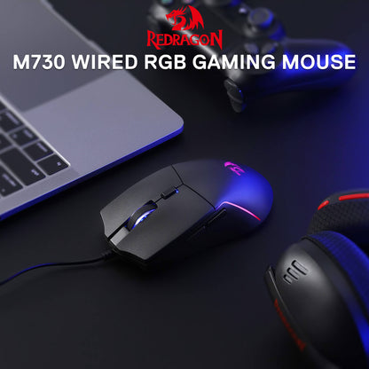 Redragon M730 wired RGB gaming mouse on a dark surface with laptop and keyboard in the background.